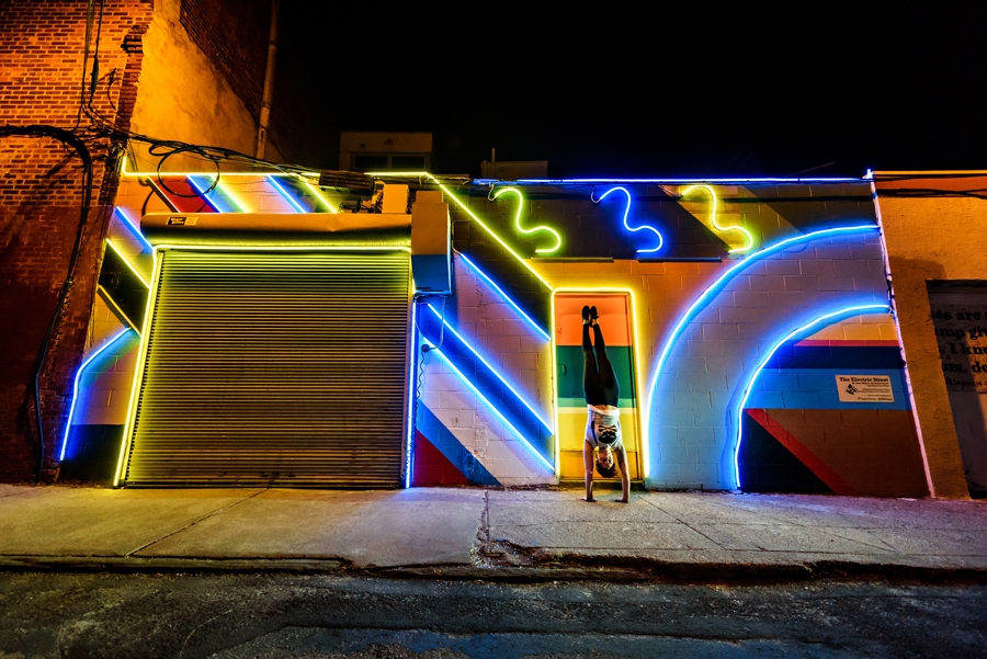 A woman doing handstands in front of beautiful neon light graffiti in south philly.